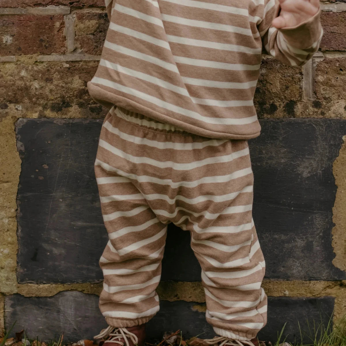 A child wearing a matching beige and cream striped long-sleeve top and striped harem pants, standing outdoors on grass in front of a brick and stone wall.
