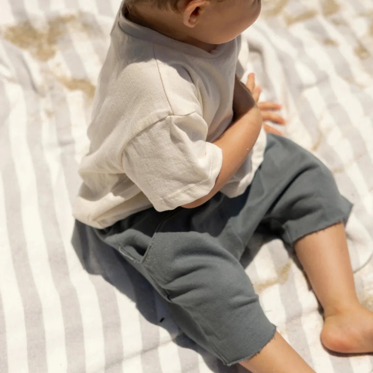 Child wearing a light gray shirt and dark gray pants sitting on a striped blanket.