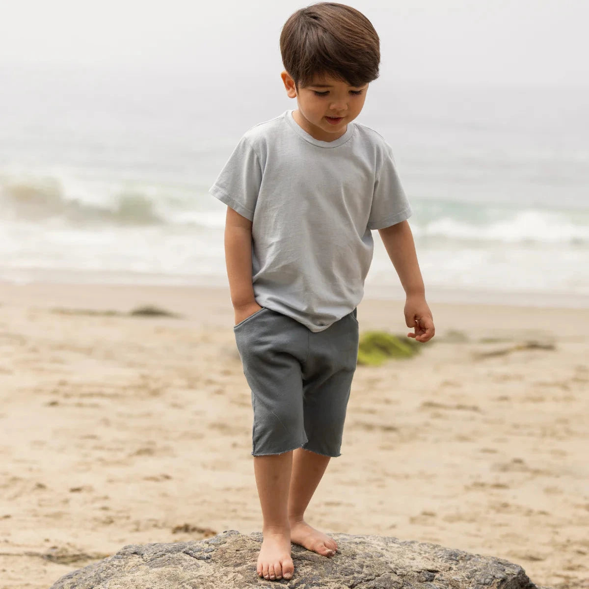 Child wearing a light gray shirt and dark gray pants sitting on a striped blanket.