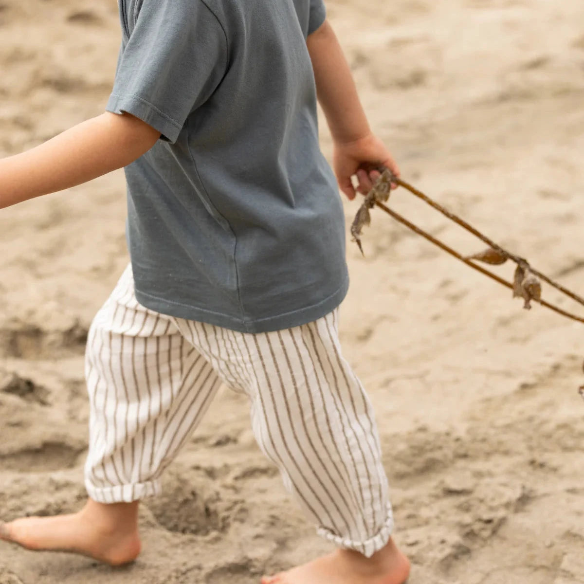 Child in gray shirt and striped pants walking on sand with a stick