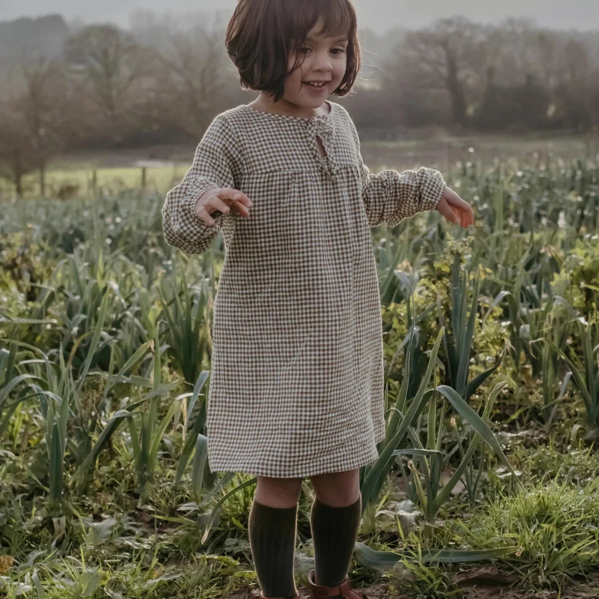 A young child wearing a beige and black gingham dress, dark green socks, and brown leather shoes stands in a grassy field.