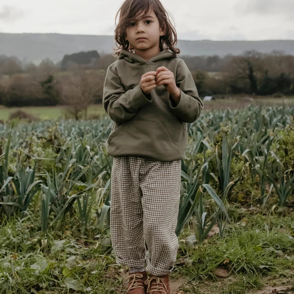 A young child standing outdoors in a field, wearing an olive green hooded sweatshirt, beige-and-white gingham pants, and brown boots.