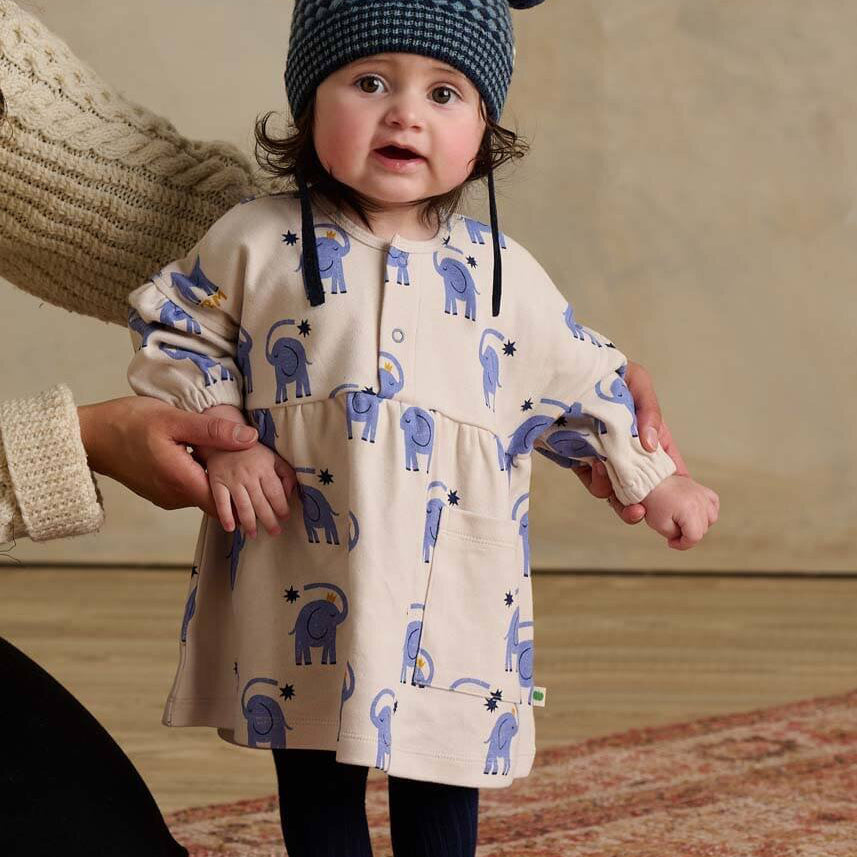 Child wearing a patterned dress and hat, standing on a wooden floor.