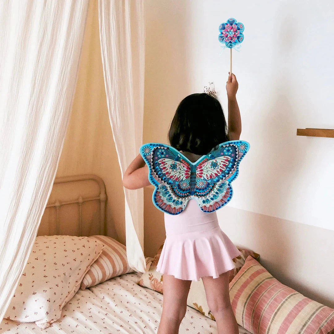 Child wearing a butterfly costume with a wand, standing on a bed in a bedroom.