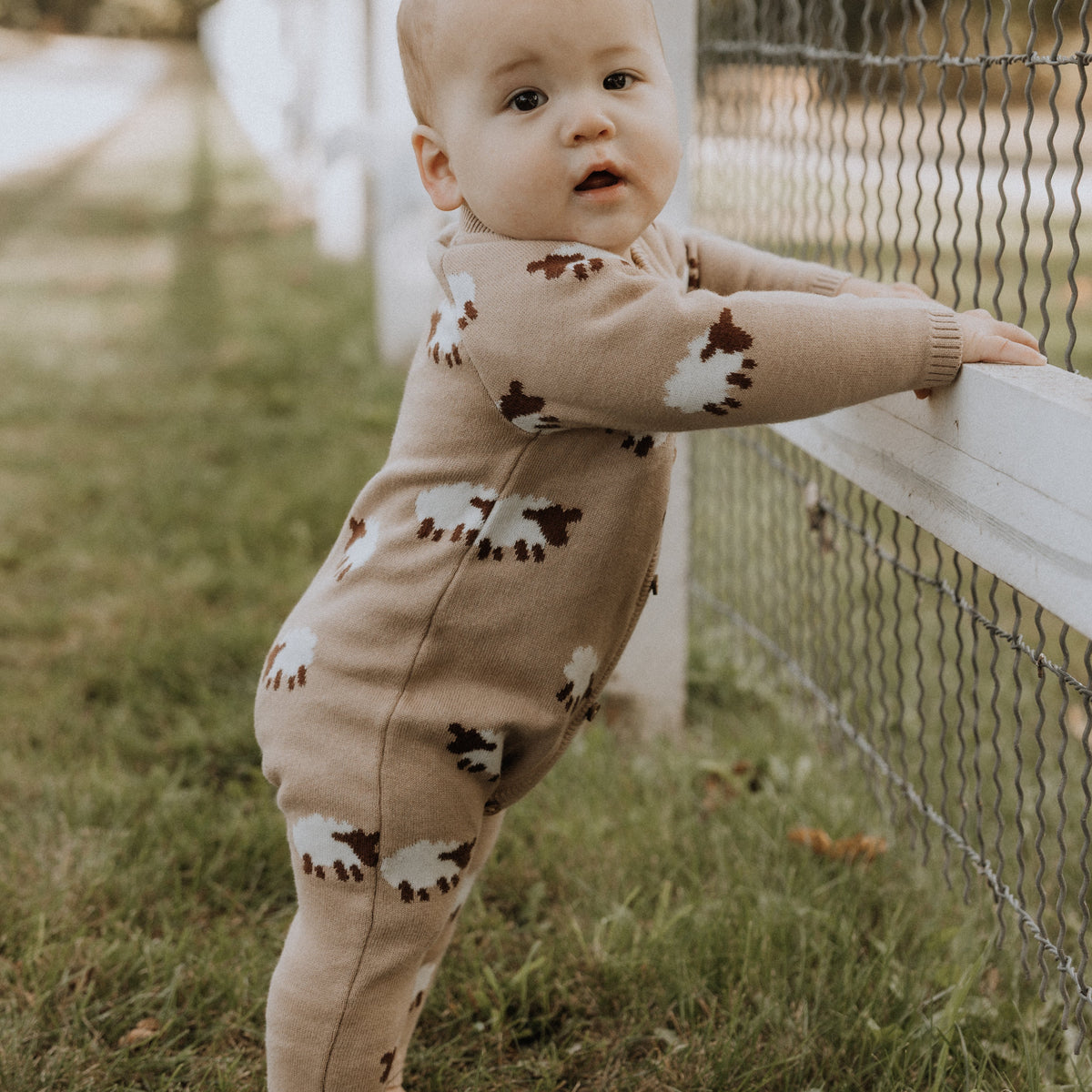 Baby in a beige romper with sheep patterns standing on grass next to a white fence.