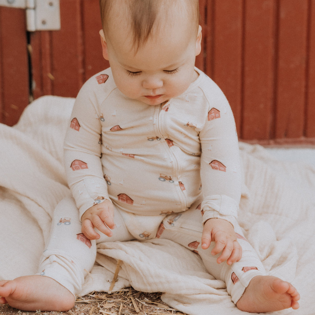 Baby sitting on straw wearing a white pajama with farm patterns against a red wooden background