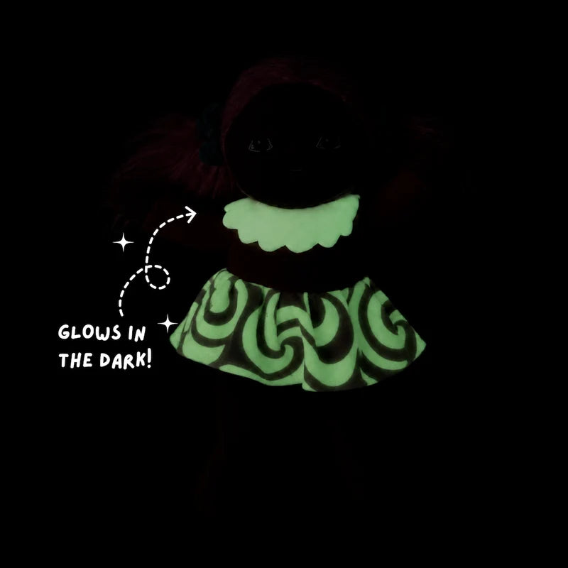 A dark, glow-in-the-dark doll shown against a black background, with a bright neon green ruffled collar and a glowing green and black patterned dress. White text on the left reads 'GLOWS IN THE DARK!' with an arrow pointing to the outfit.