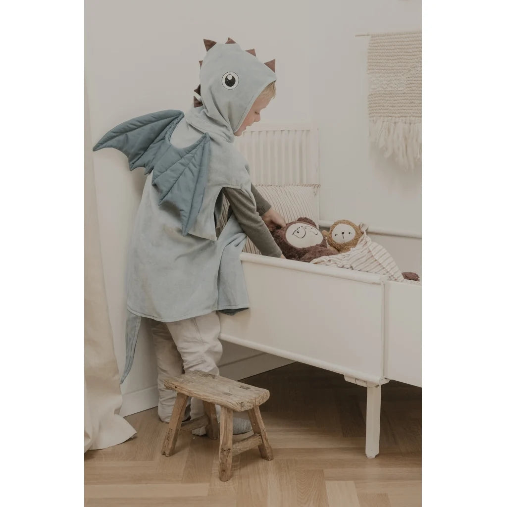 A child wearing a light blue-gray dino hooded cape with fabric wings and a tail, standing on a small wooden stool beside a white children's bed in a softly lit nursery.