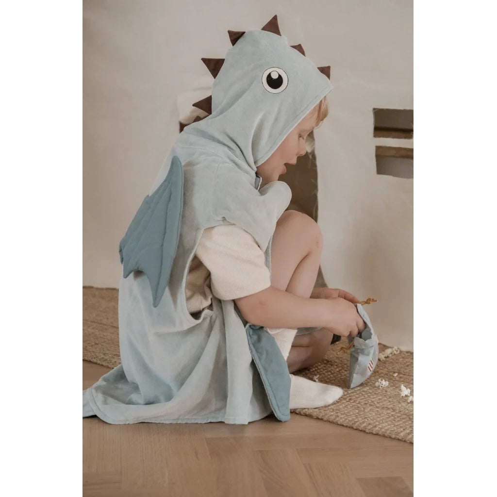 A child wearing a light blue dinosaur hooded towel cape, sitting on a woven rug on a wooden floor.