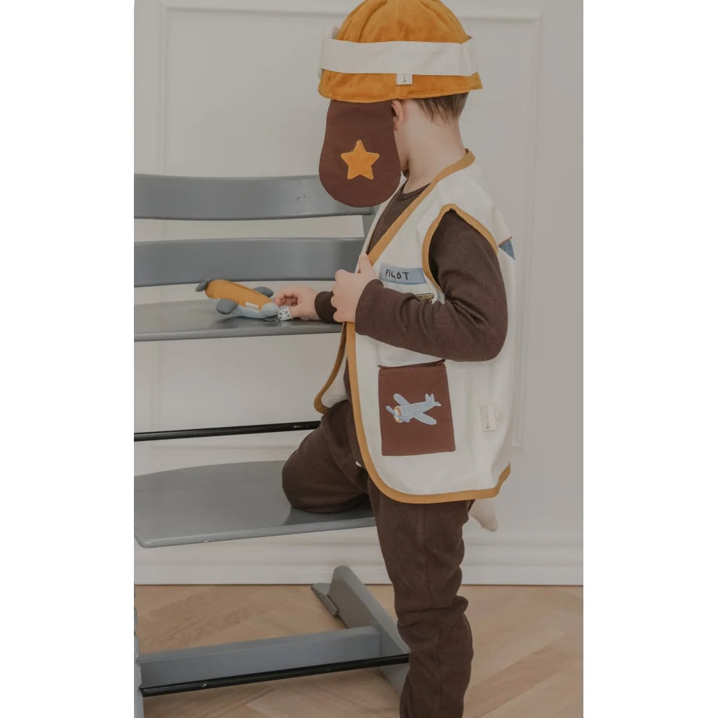A child wearing a brown and cream airplane-themed costume, including a matching hat, stands next to a gray wooden step stool in an indoor room.