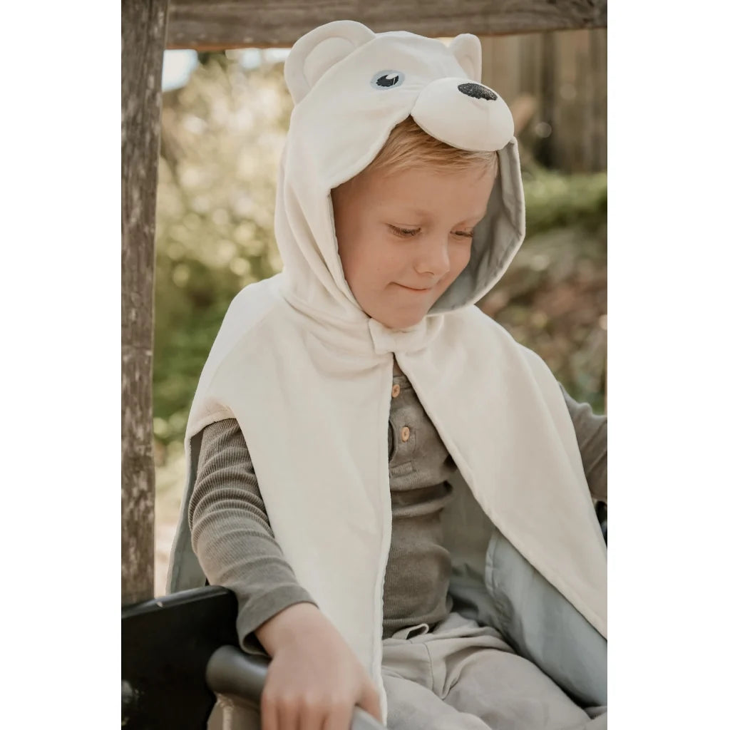 A young child wearing a cream-colored fleece hooded cape with a bear face design on the hood, sitting outdoors.