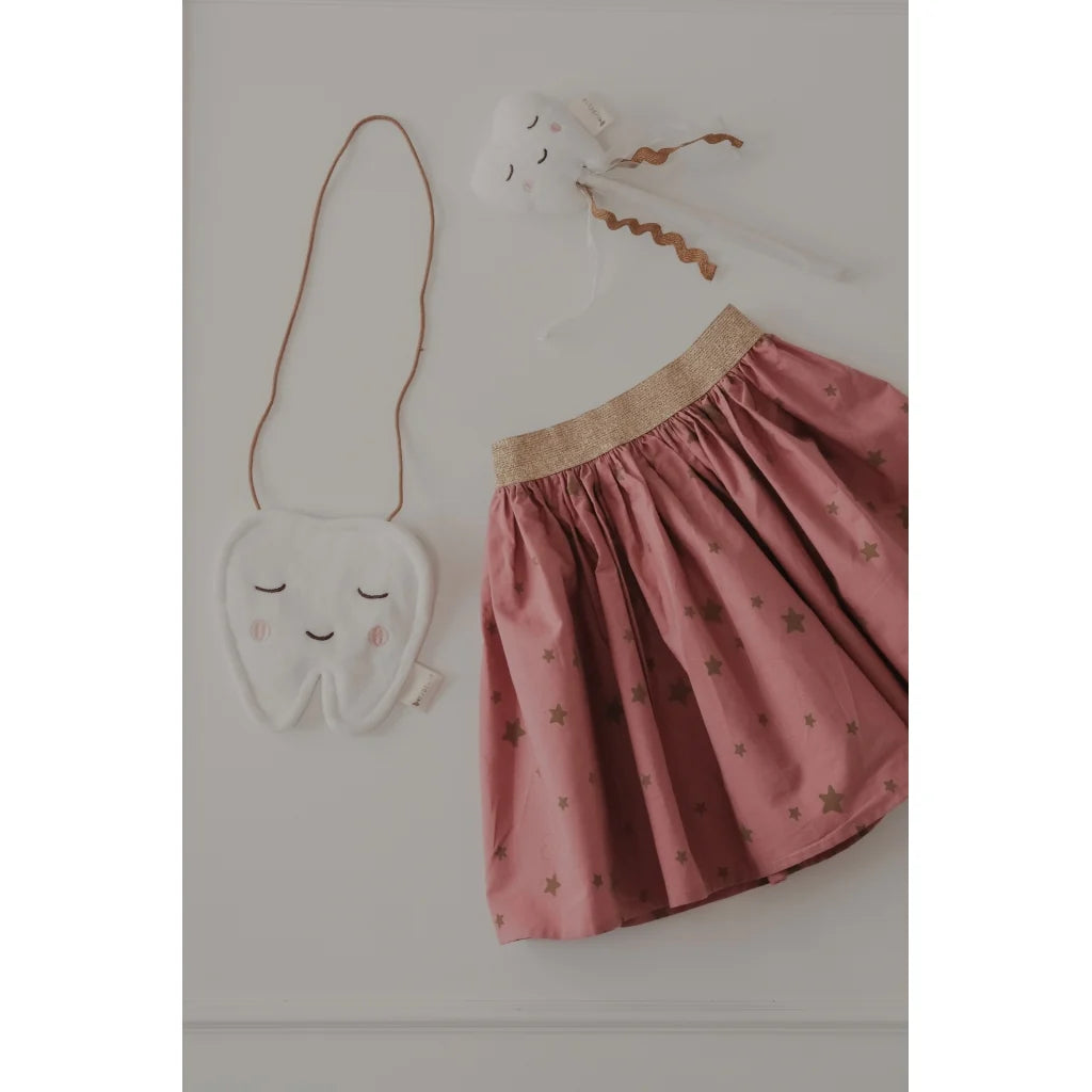 A children's outfit set featuring a dusty pink skirt with small gold star prints and a beige elastic waistband, styled with a white tooth-shaped shoulder bag and a white bunny head accessory on a light gray background.