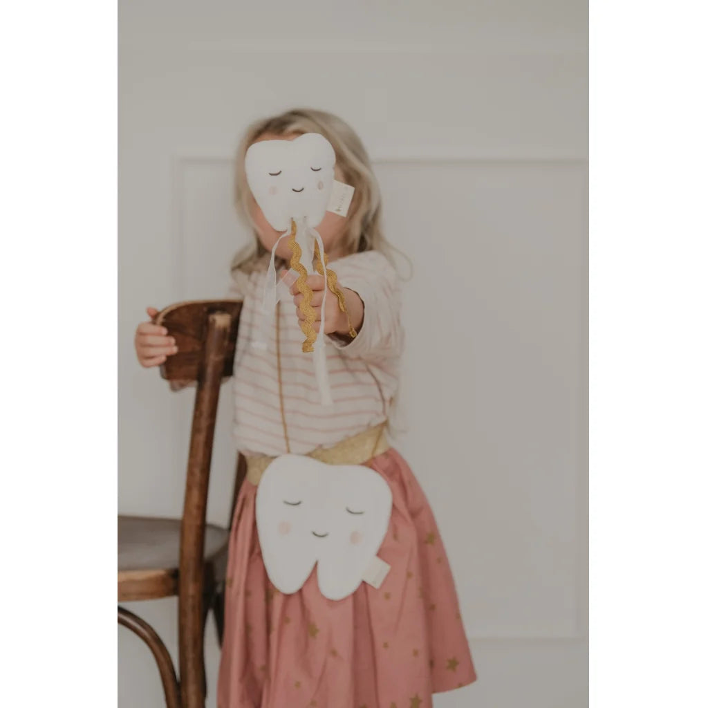 A young child wearing a pink skirt with gold stars and a striped top holds a white tooth-shaped wand  while wearing a matching white tooth-shaped crossbody bag with a gold strap.