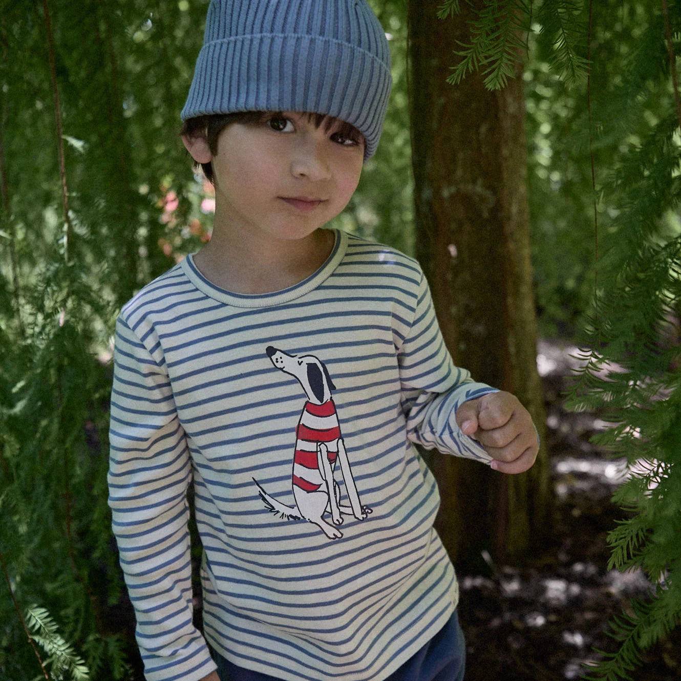 Child wearing a blue beanie and striped shirt with a dog graphic in a forest setting