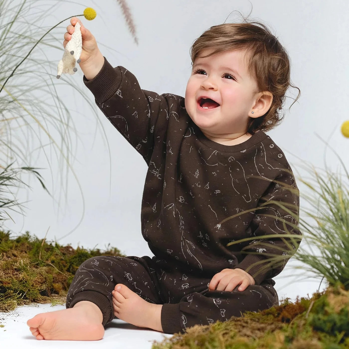 Child in a brown outfit sitting on grass holding a flower