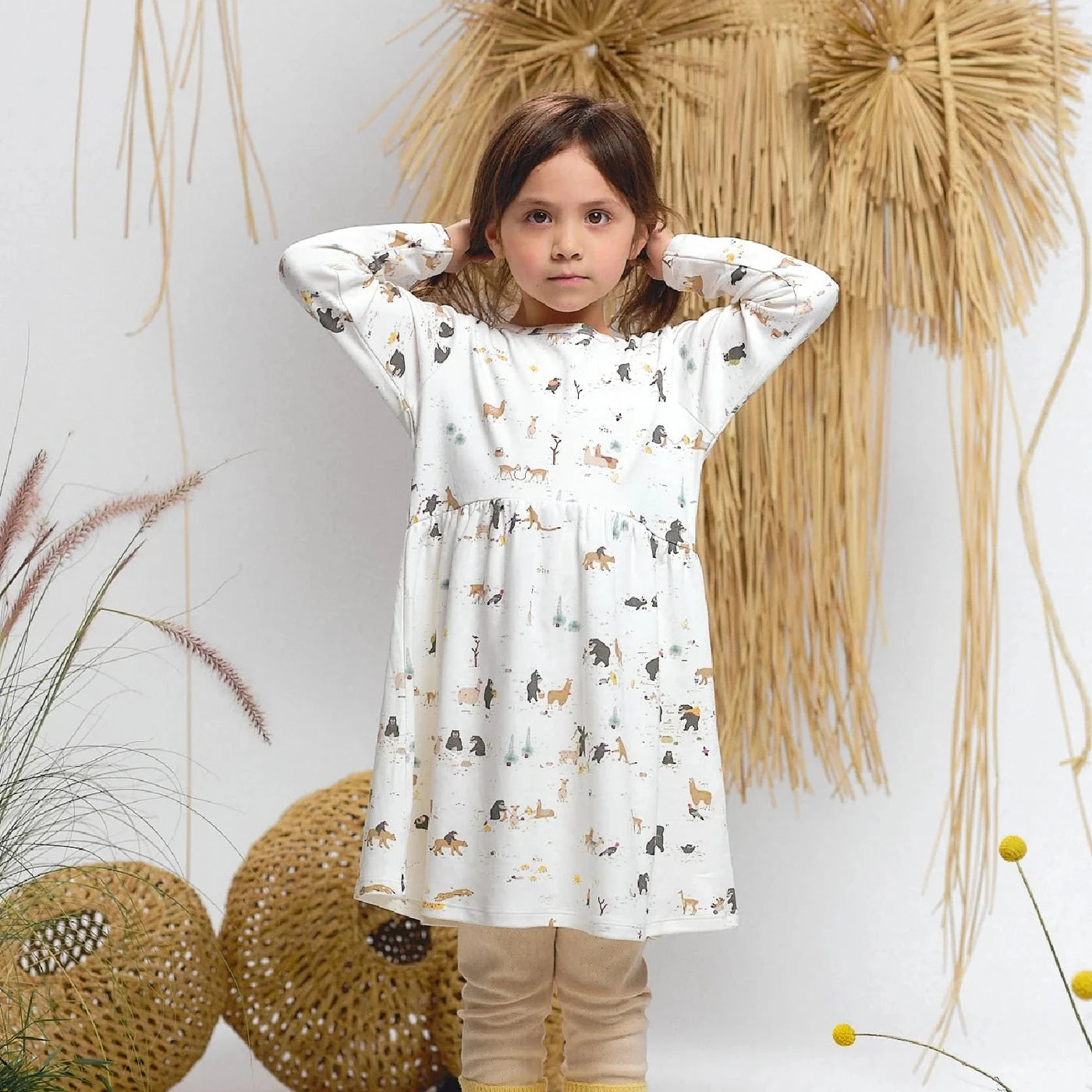 Young girl wearing a patterned dress with yellow leggings standing in front of decorative plants.
