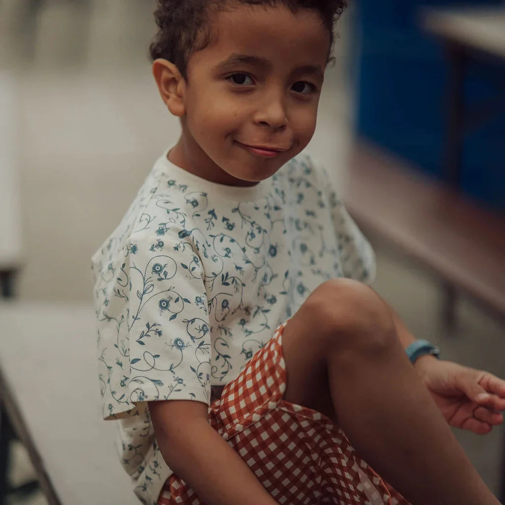 Child sitting on a bench wearing a floral shirt and checkered shorts.
