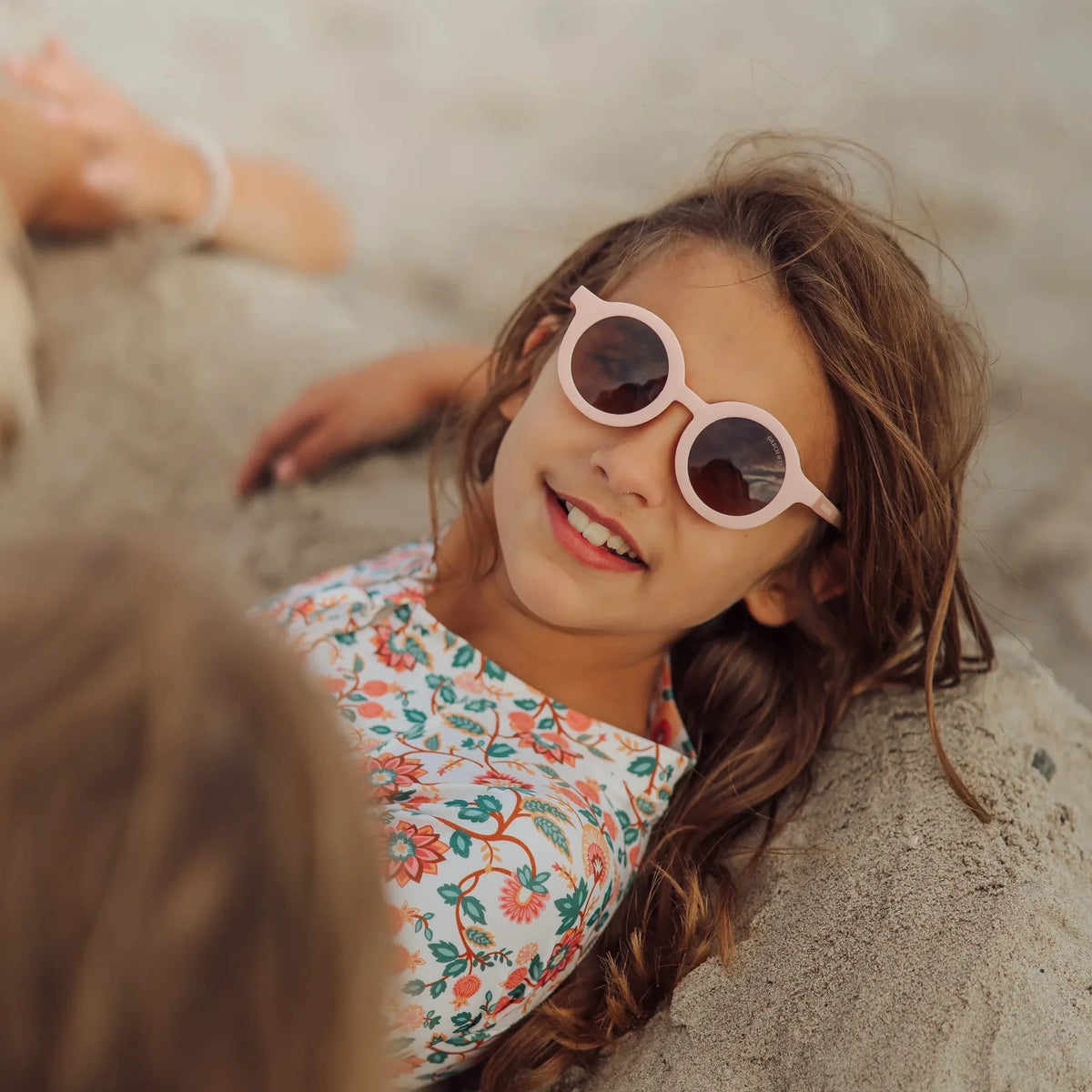 Child wearing sunglasses and a floral dress lying on sand