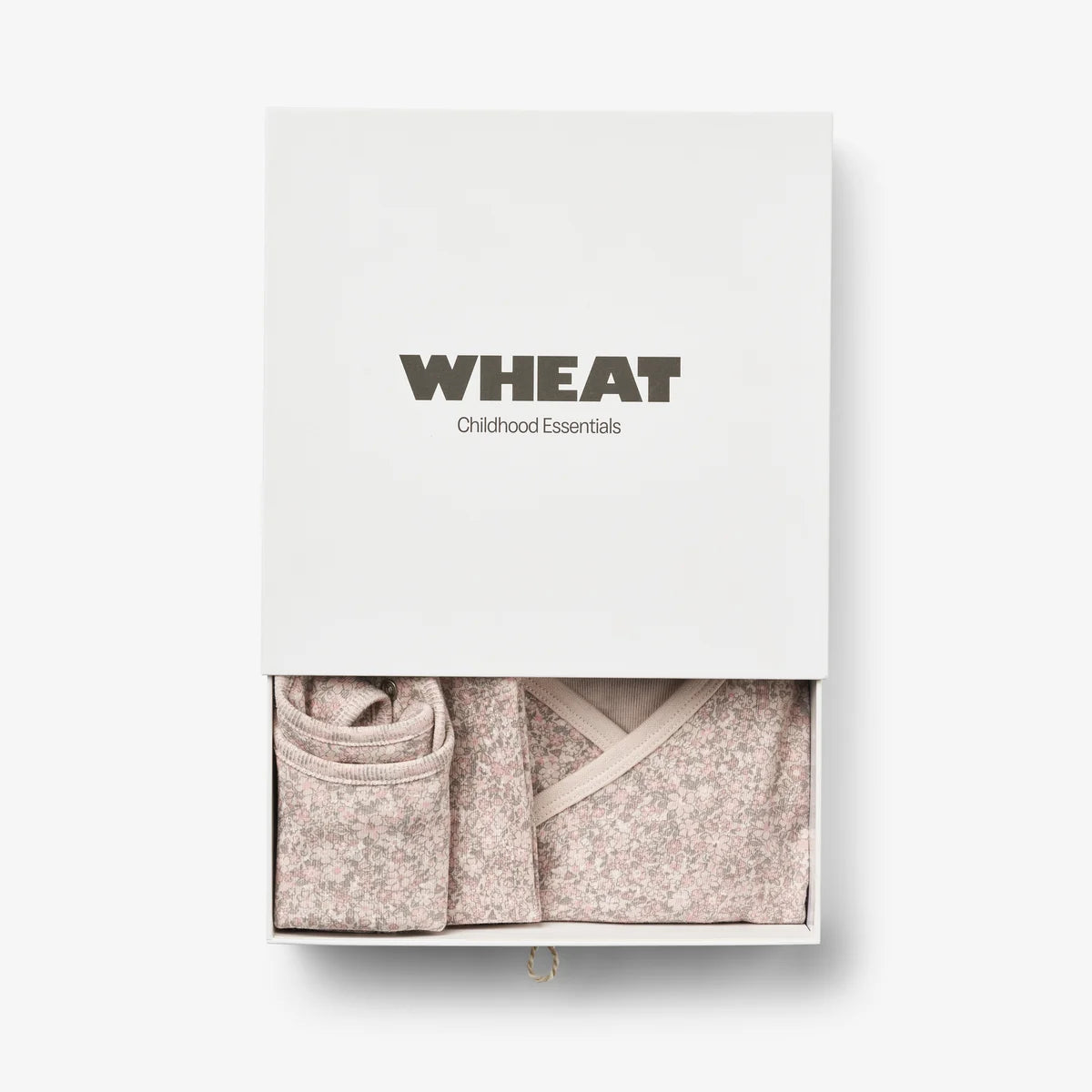 A white gift box labeled 'WHEAT Childhood Essentials' with a sliding top section open to reveal folded knit baby clothing in a soft blush-beige speckled fabric, set against a clean white background.