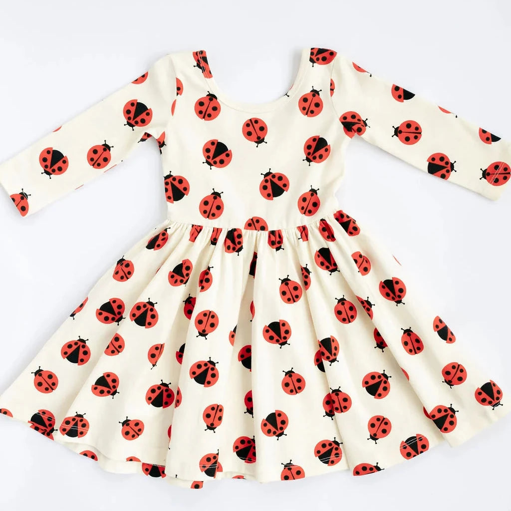 A long-sleeve dress with a flared skirt, featuring an all-over red ladybug print on a warm cream background, laid flat against a white background.
