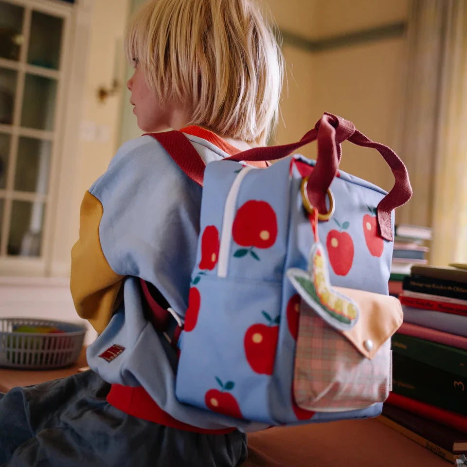 Child wearing a blue backpack with red apple patterns in a room.