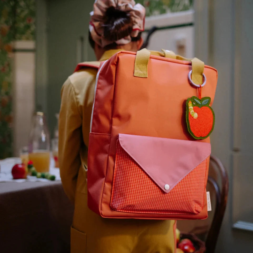 Person wearing an orange backpack with a green apple charm in a room with floral wallpaper.