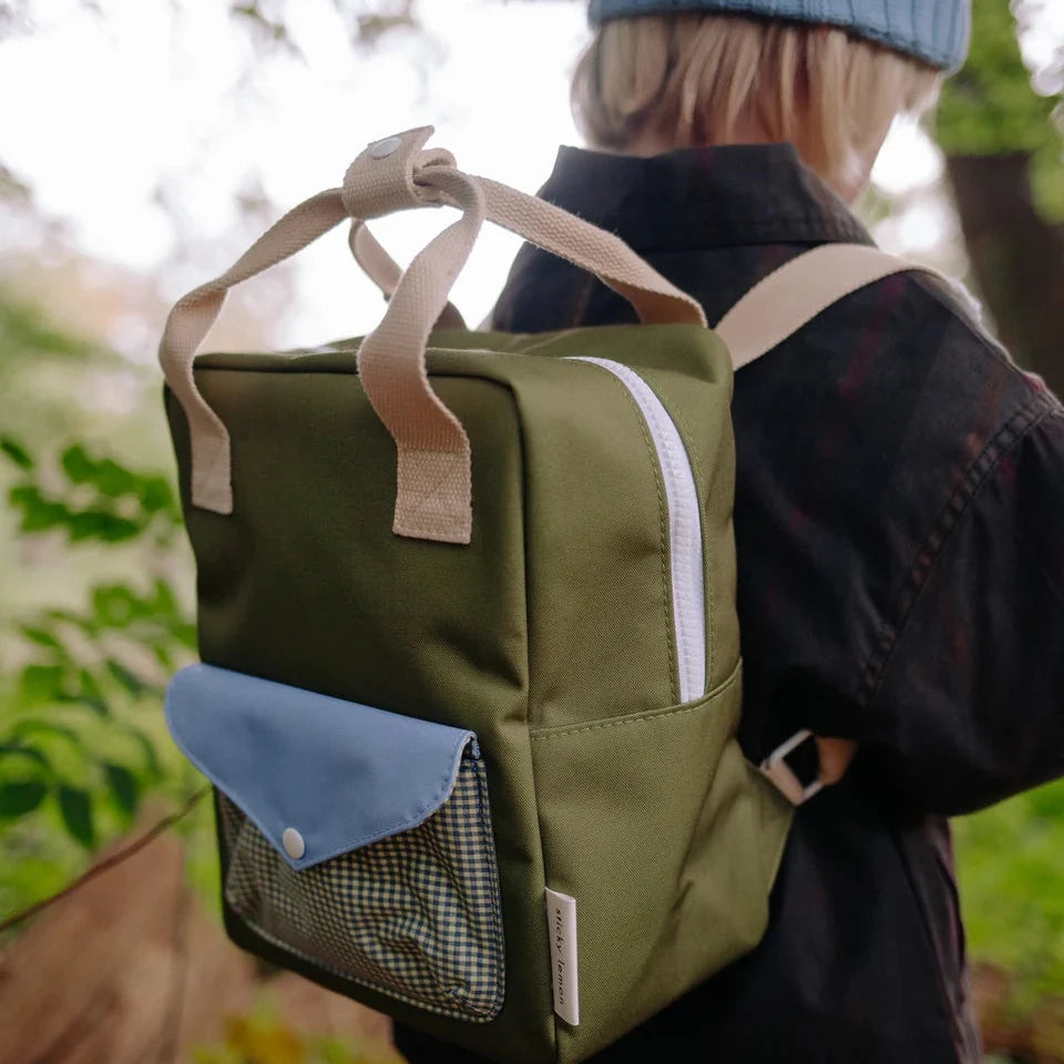 An olive green canvas backpack with beige handles and a light blue front pocket, shown from behind on a child wearing a beanie and dark jacket in an outdoor setting.