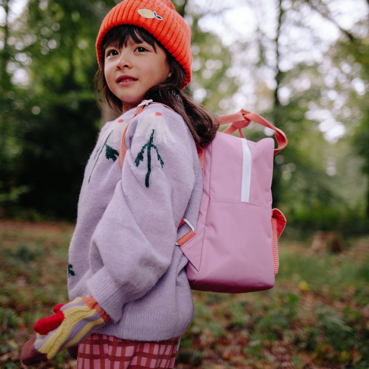 Child wearing a pink backpack and orange beanie in a forest setting