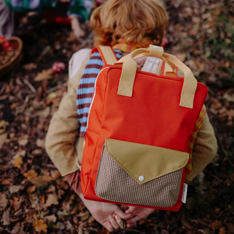 Person wearing a red backpack with a yellow flap in an outdoor setting with leaves on the ground.