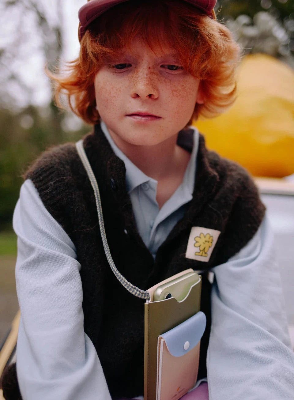 Young boy with red hair wearing a cap and jacket, holding a book outdoors.
