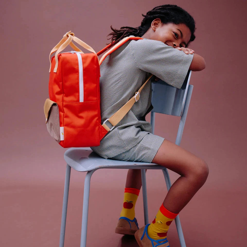 Person sitting on a chair with an orange backpack, wearing colorful socks and shoes against a brown background