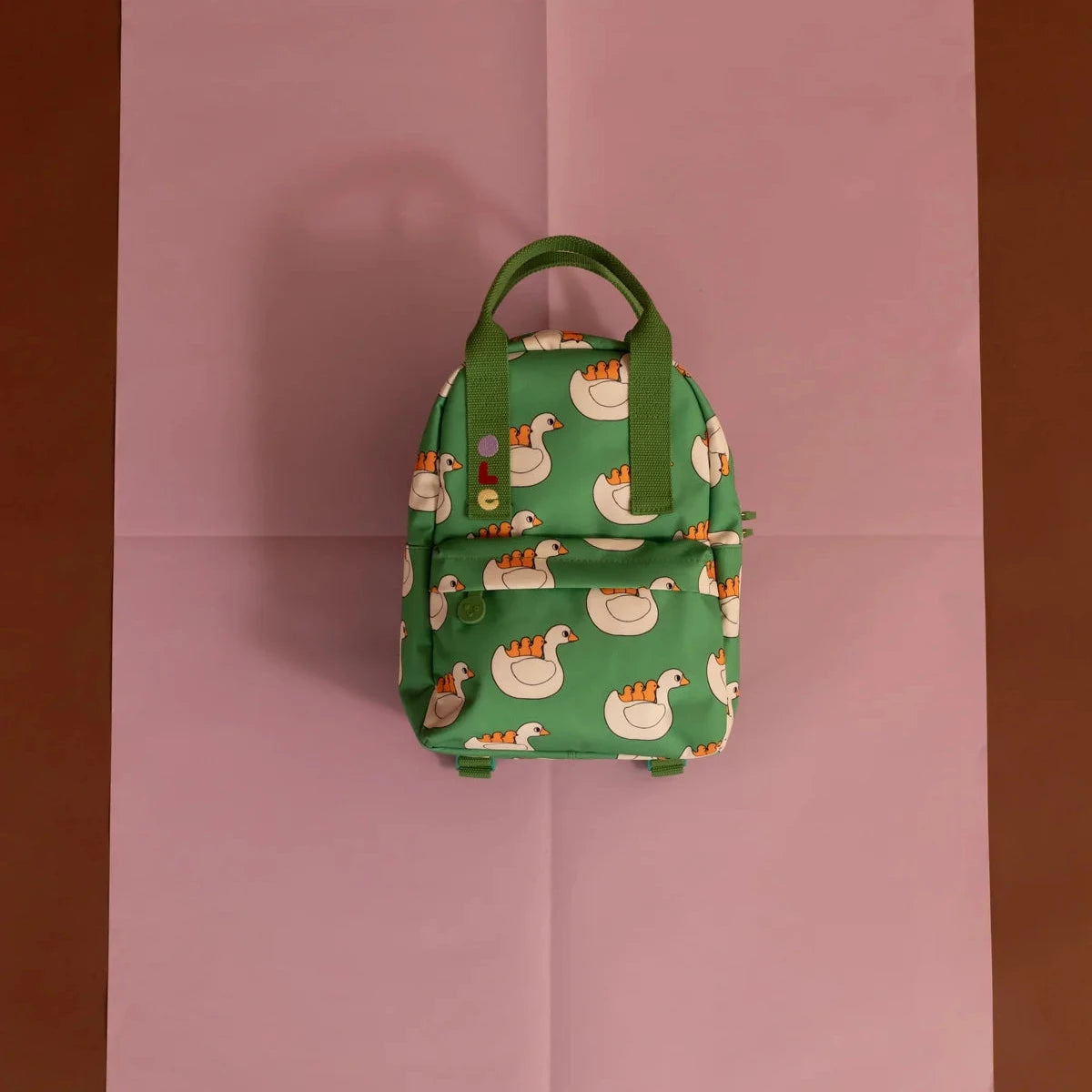 A green backpack with a white swan pattern, featuring green handles and a front zipper pocket, centered on a pink sheet of paper against a brown background.