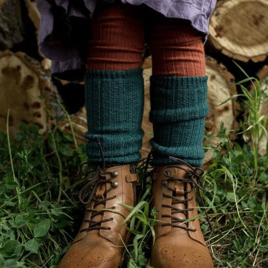 Child wearing brown boots and green leg warmers standing in grass with wooden logs in the background