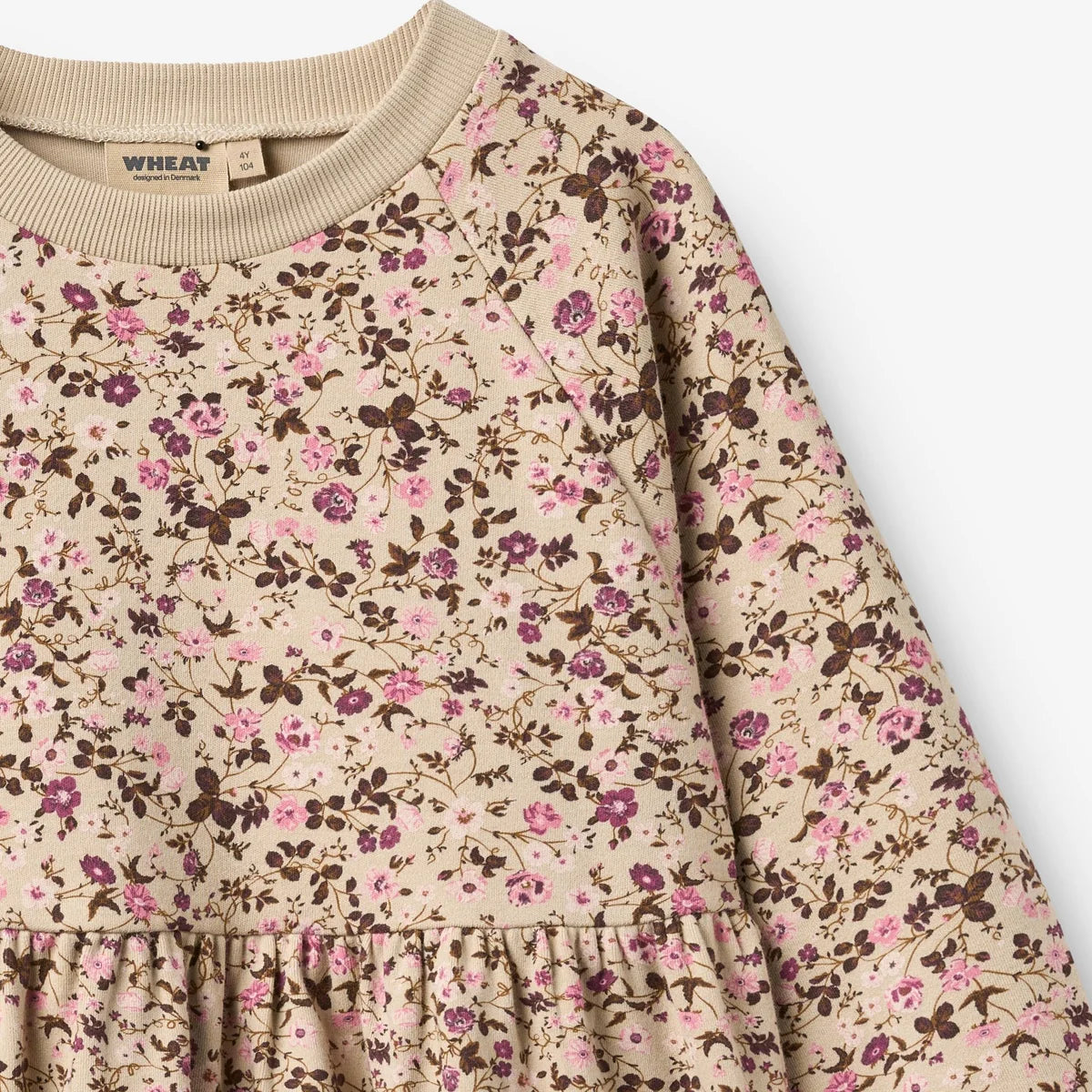 Close-up of a long-sleeve children's dress with a beige crew neck and an all-over floral print in pink, purple, and brown on a cream background. The brand name 'WHEAT' is visible on the inner neck label.