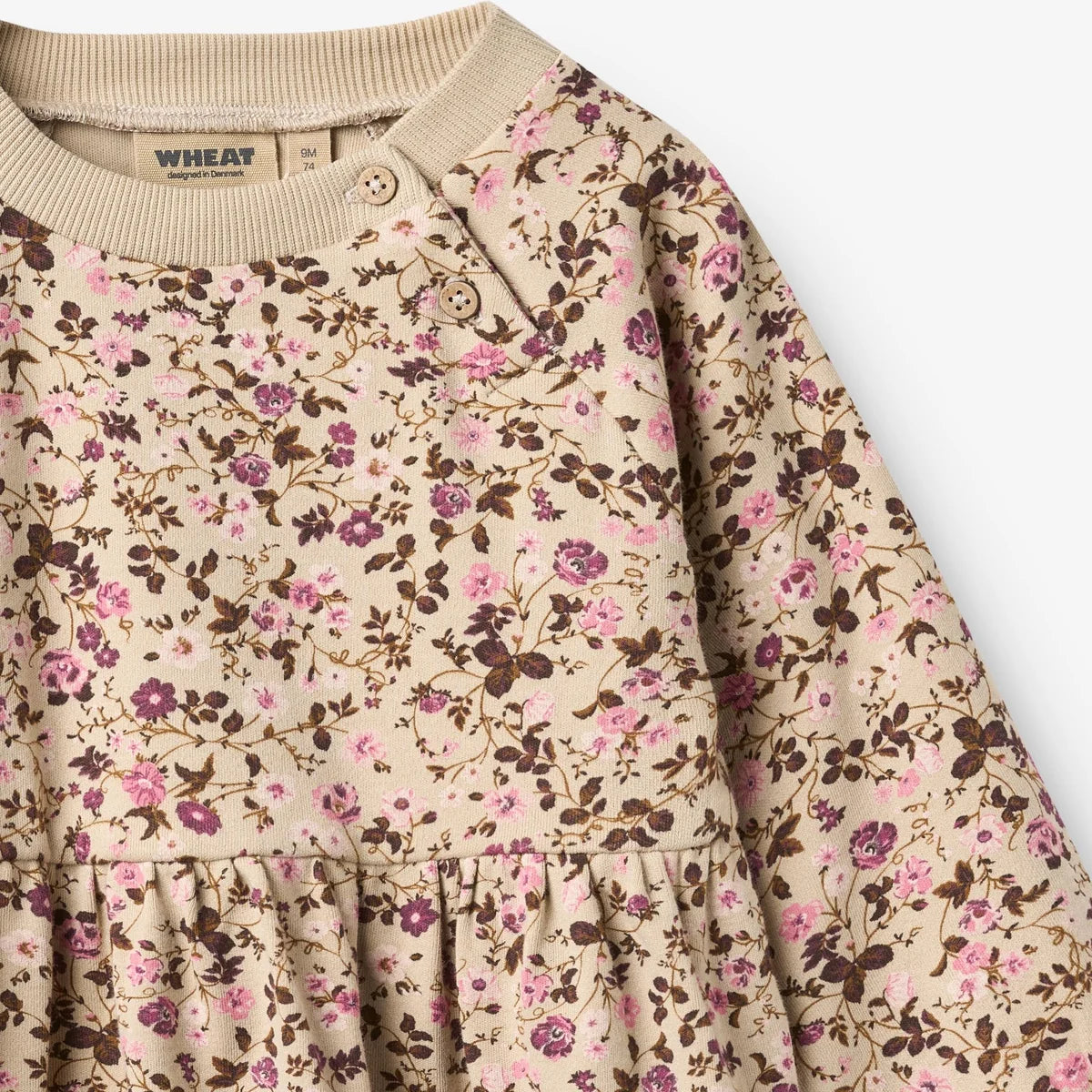 Close-up of a baby's long-sleeve dress with a beige crew neck and an all-over floral print in pink, purple, and brown on a cream background. The brand name 'WHEAT' is visible on the inner neck label.