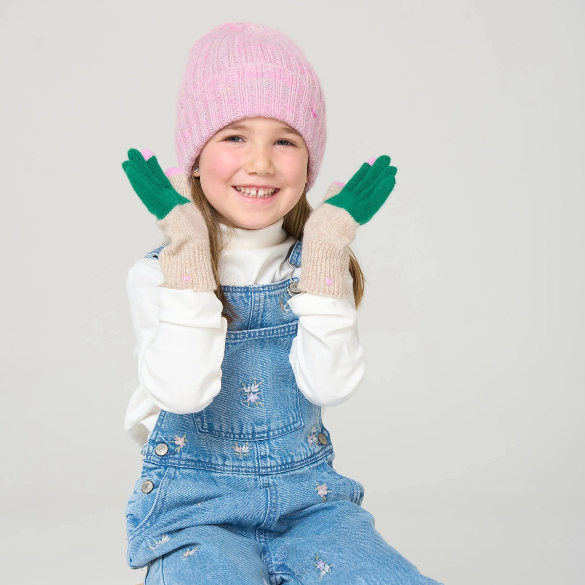 Child wearing a pink knit hat, white gloves, and blue denim overalls sitting on a wooden stool against a light gray background.
