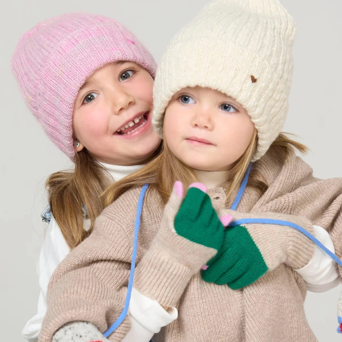 Child wearing a pink knit hat, white gloves, and blue denim overalls sitting on a wooden stool against a light gray background.