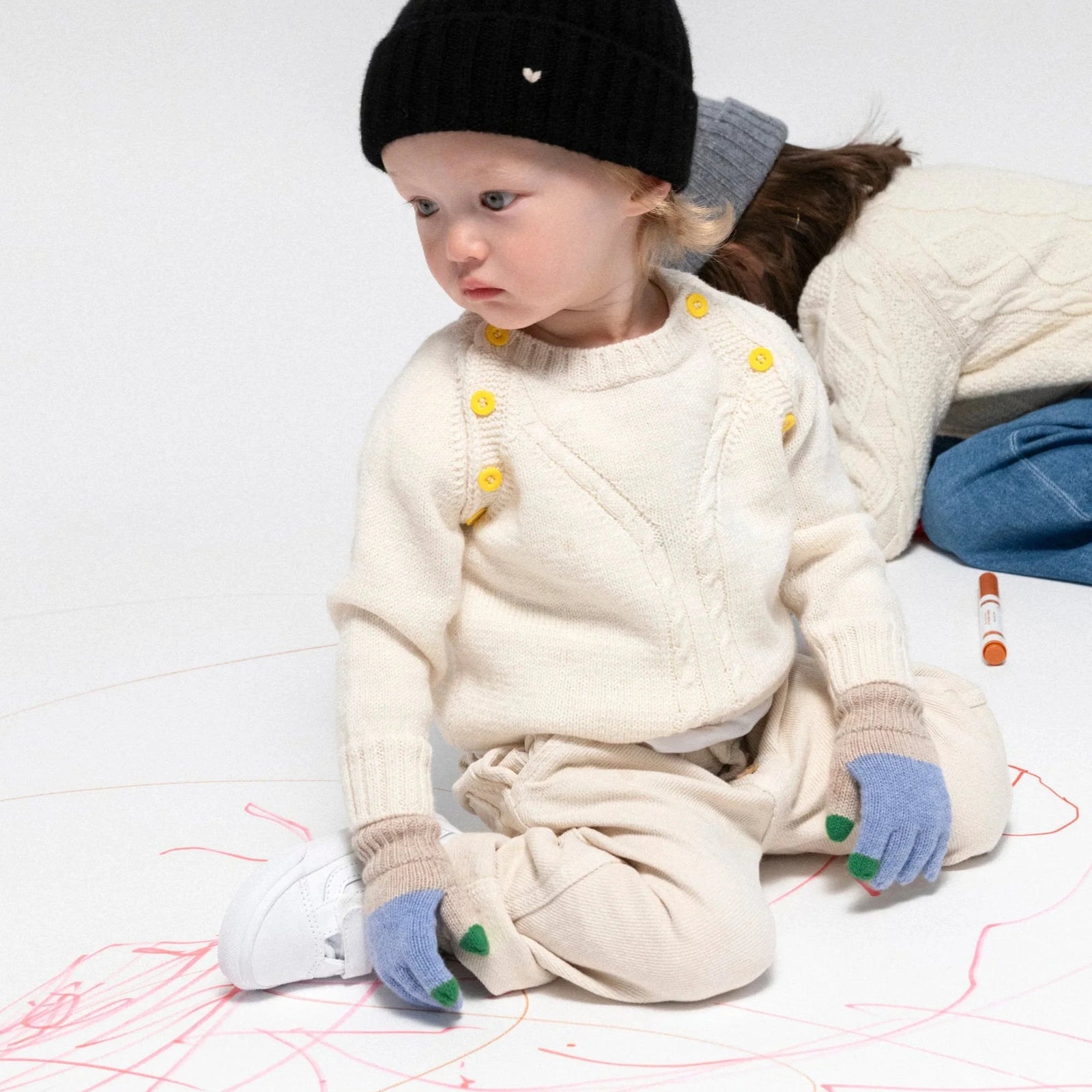 Child wearing a pink knit hat, white gloves, and blue denim overalls sitting on a wooden stool against a light gray background.