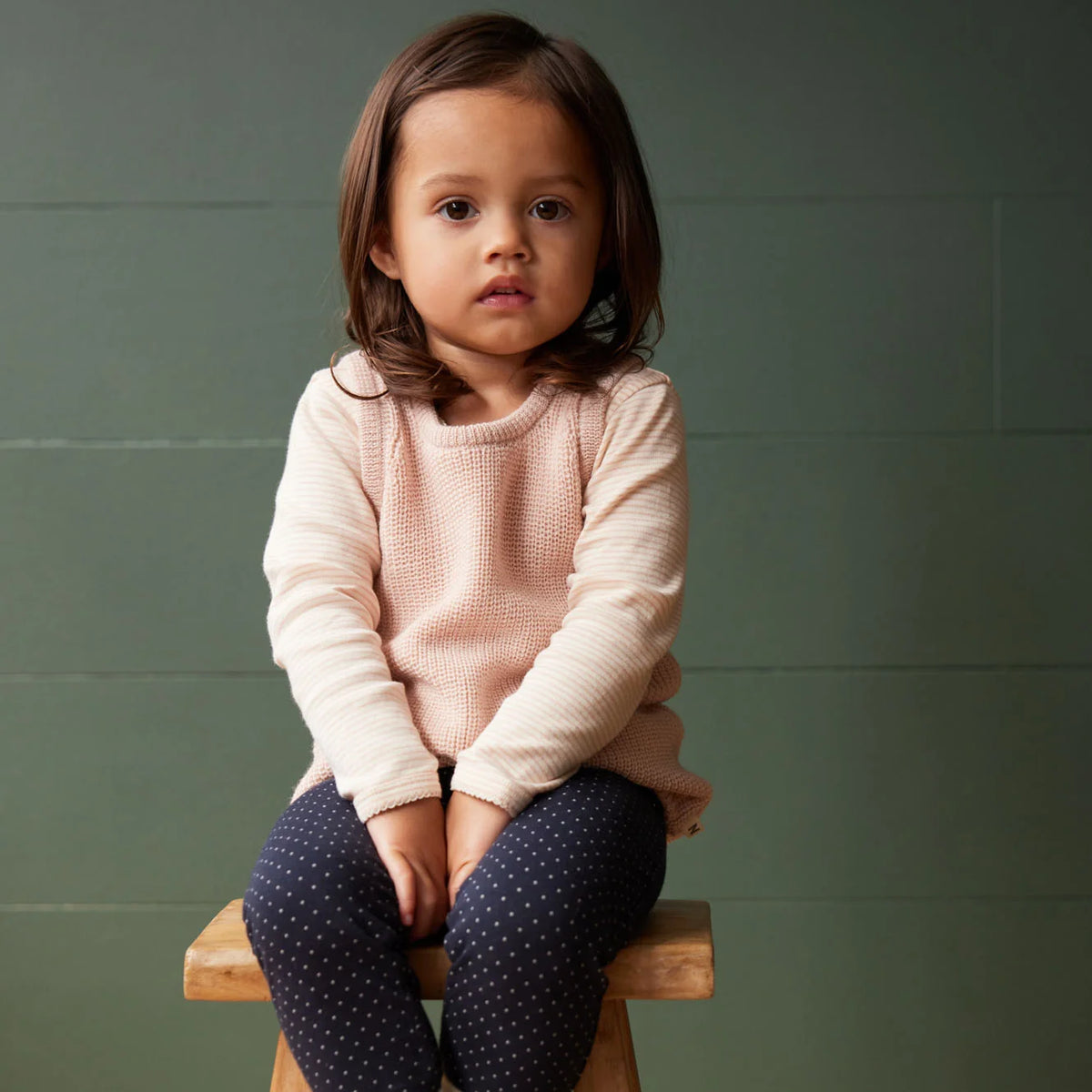 A young child with brown hair sits on a wooden stool against a muted green wall, wearing a pale pink textured knit sweater and navy blue leggings with small white polka dots.