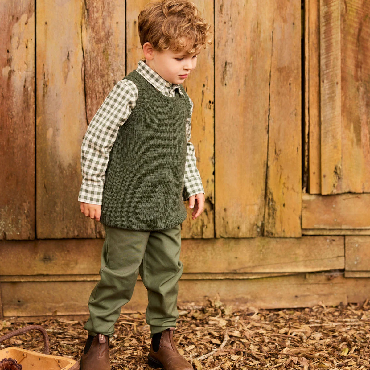A young boy wearing an olive green knit sweater vest over a green and white gingham shirt, olive green pants, and brown boots, standing in front of a rustic wooden fence.