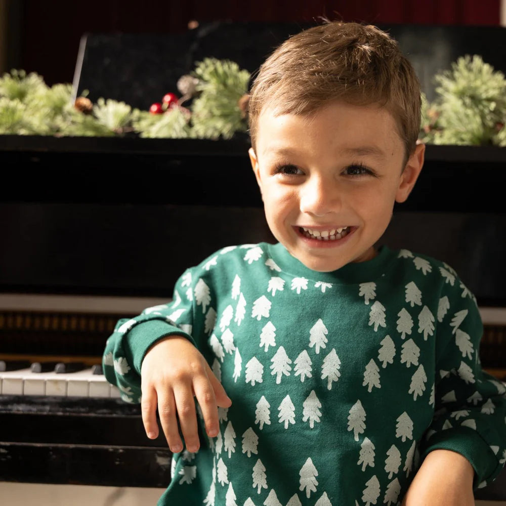 Child wearing a green sweater with white tree patterns, sitting in front of a piano.