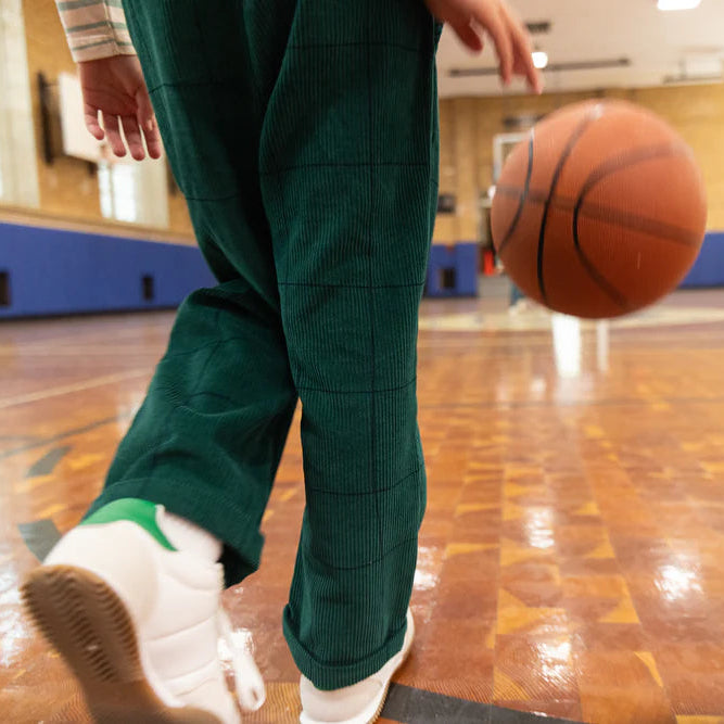 Person in green pants and white shoes dribbling a basketball on a gymnasium floor.