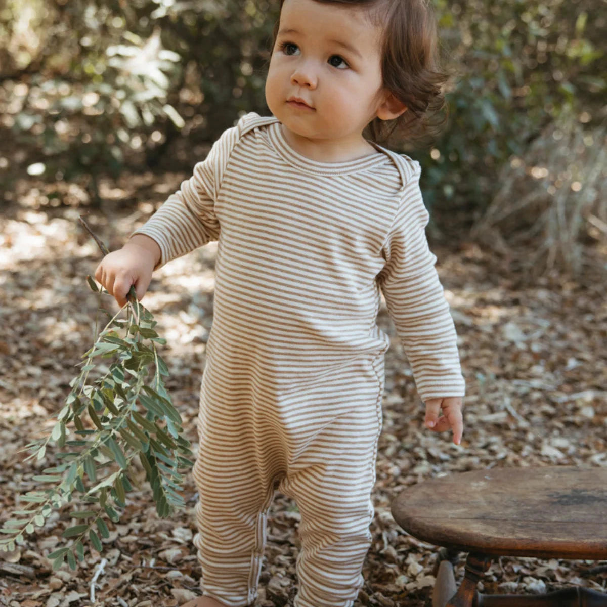 Child in a striped romper standing outdoors with leaves in hand