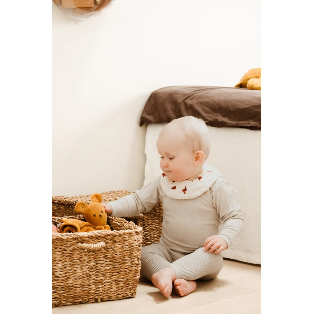 Baby playing with a teddy bear in a wicker basket on a light-colored floor.