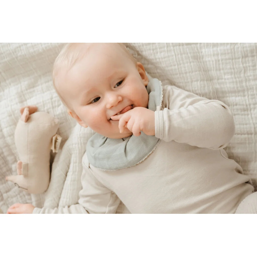 Baby lying on a textured blanket with a soft toy, wearing a light-colored outfit and a bib.