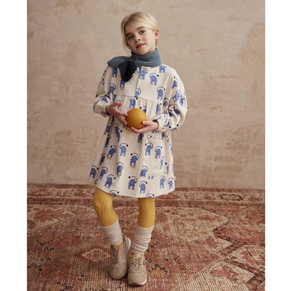 Child wearing a patterned dress and hat, standing on a wooden floor.