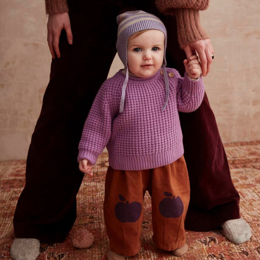 Baby in a purple sweater and brown pants standing next to an adult on a patterned rug.