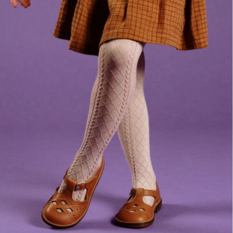 A child wearing a brown plaid skirt, cream diamond-knit tights, and brown leather mule shoes against a solid purple studio background.