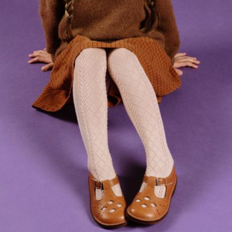 A young girl with braided hair sits on a purple studio background wearing a brown knit sweater and skirt, cream tights with a diamond pattern, and brown Mary Jane shoes.