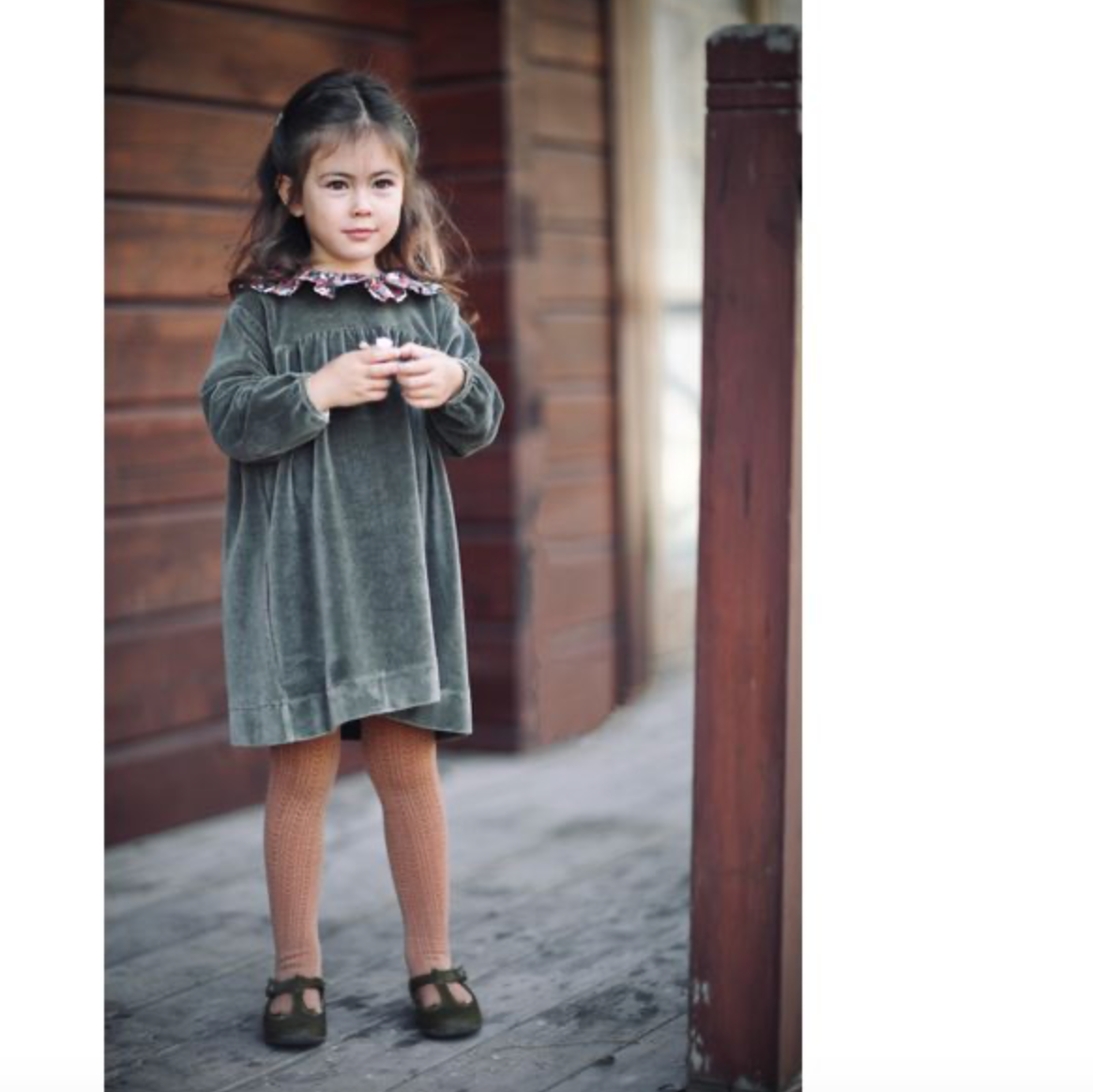 A young girl wearing a dark green velvet dress with a ruffled floral collar, brown tights, and dark brown shoes, standing on a wooden porch.